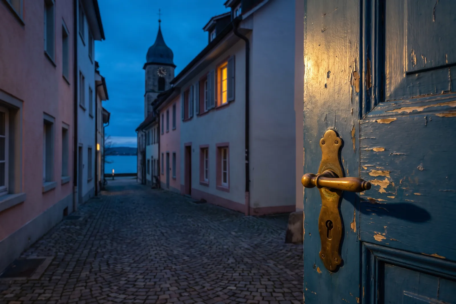 Symbolbild: verwitterte blaue Haustür mit Messingbeschlag in einer Altstadtgasse mit Blick auf Kirchturm und See – Einsatzgebiet Schlüsseldienst Radolfzell